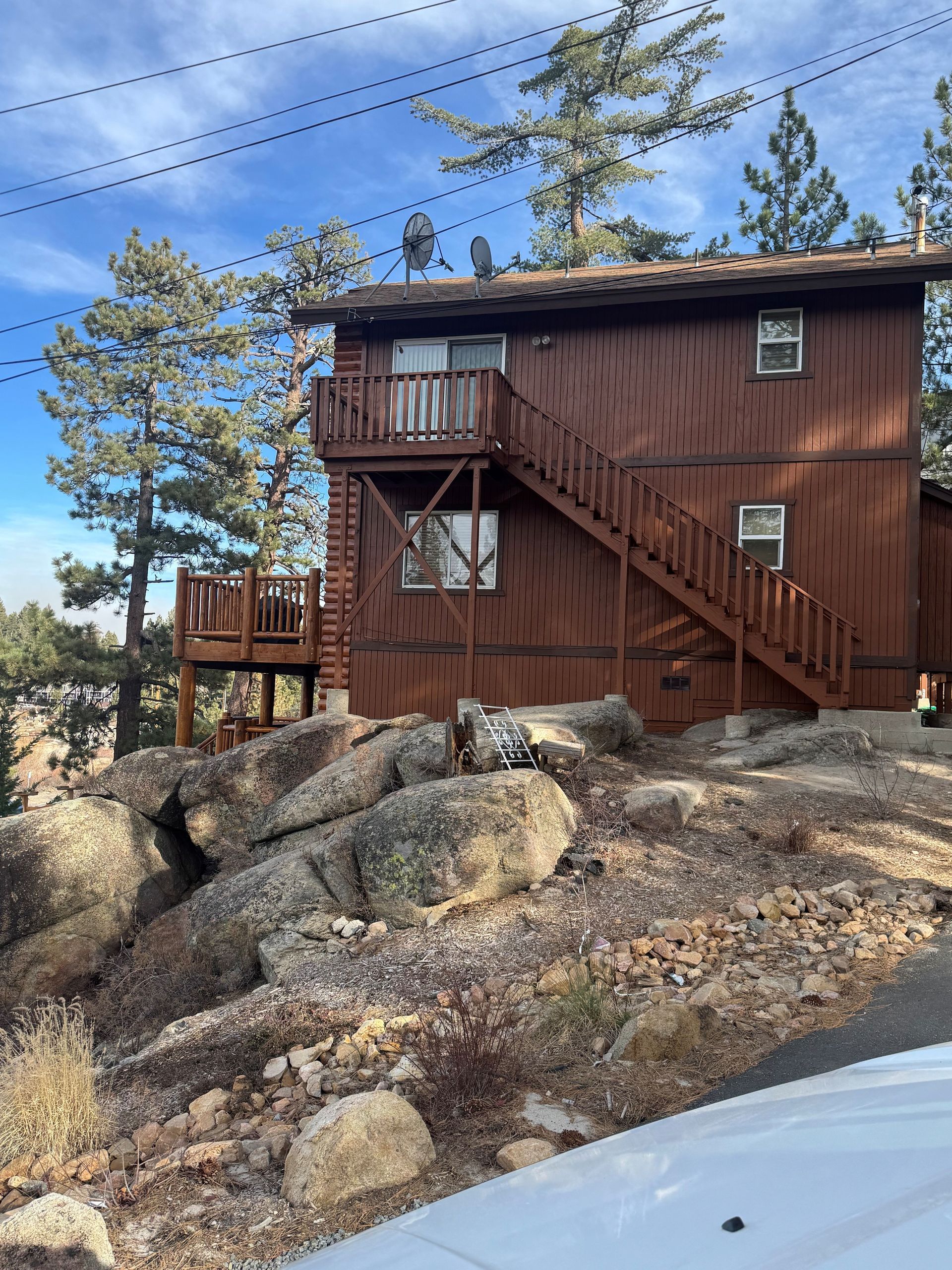 Brown cabin with exterior stairs and deck, nestled among boulders and pine trees, on a hillside.