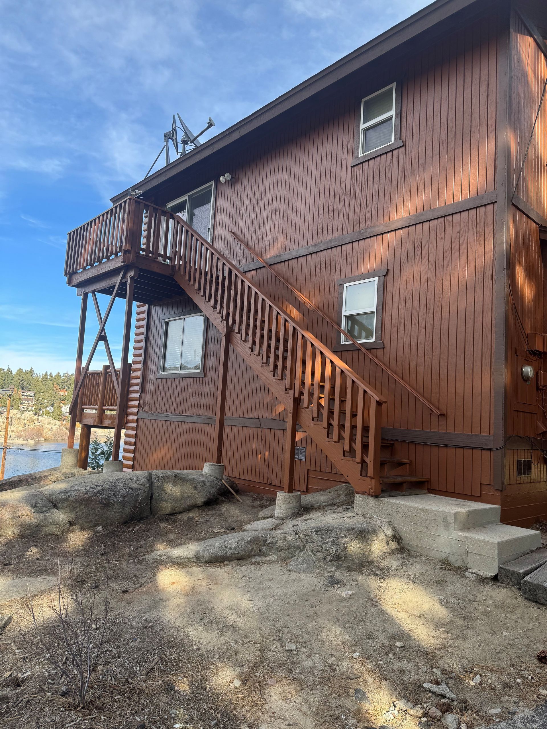 Brown two-story wooden cabin with outdoor stairs and a deck overlooking a lake, on a sunny day.