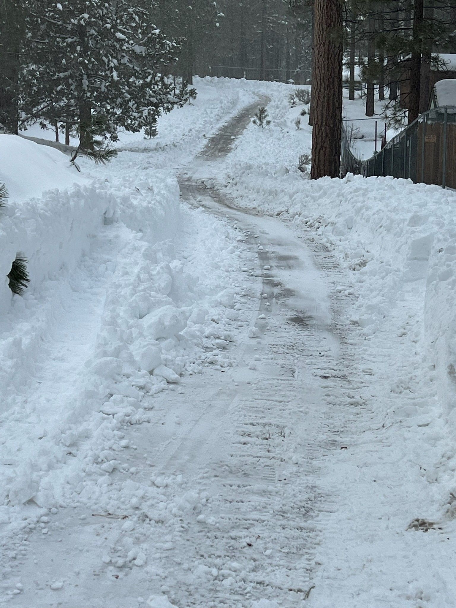 Snow-covered road carved through deep snow banks. Trees and a fence line the sides.