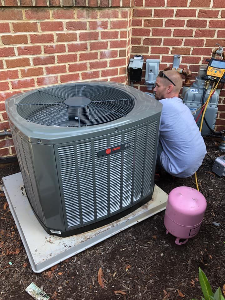 Technician working on a gray HVAC unit beside a brick wall, with tools and a pink tank nearby.