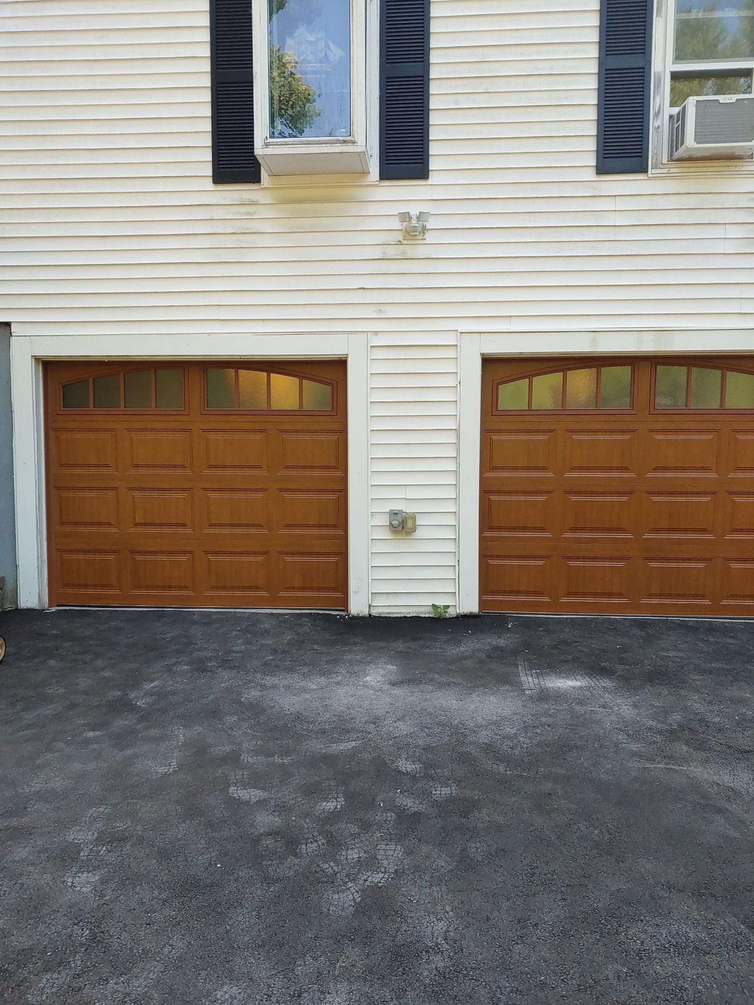 Two brown garage doors on a weathered white house. Black asphalt driveway.