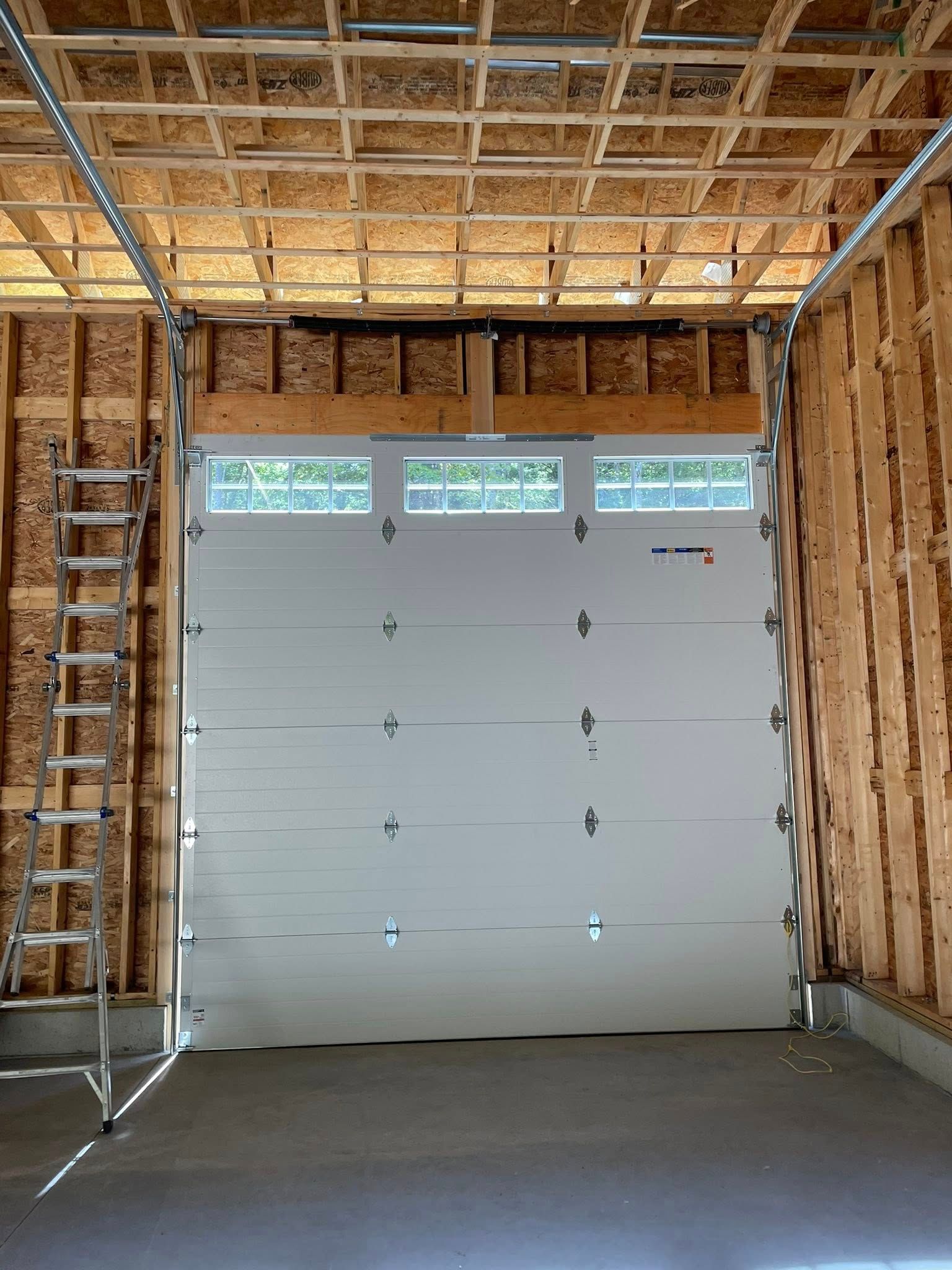 Garage door, white, installed in an unfinished wooden garage with visible framing and ceiling beams.