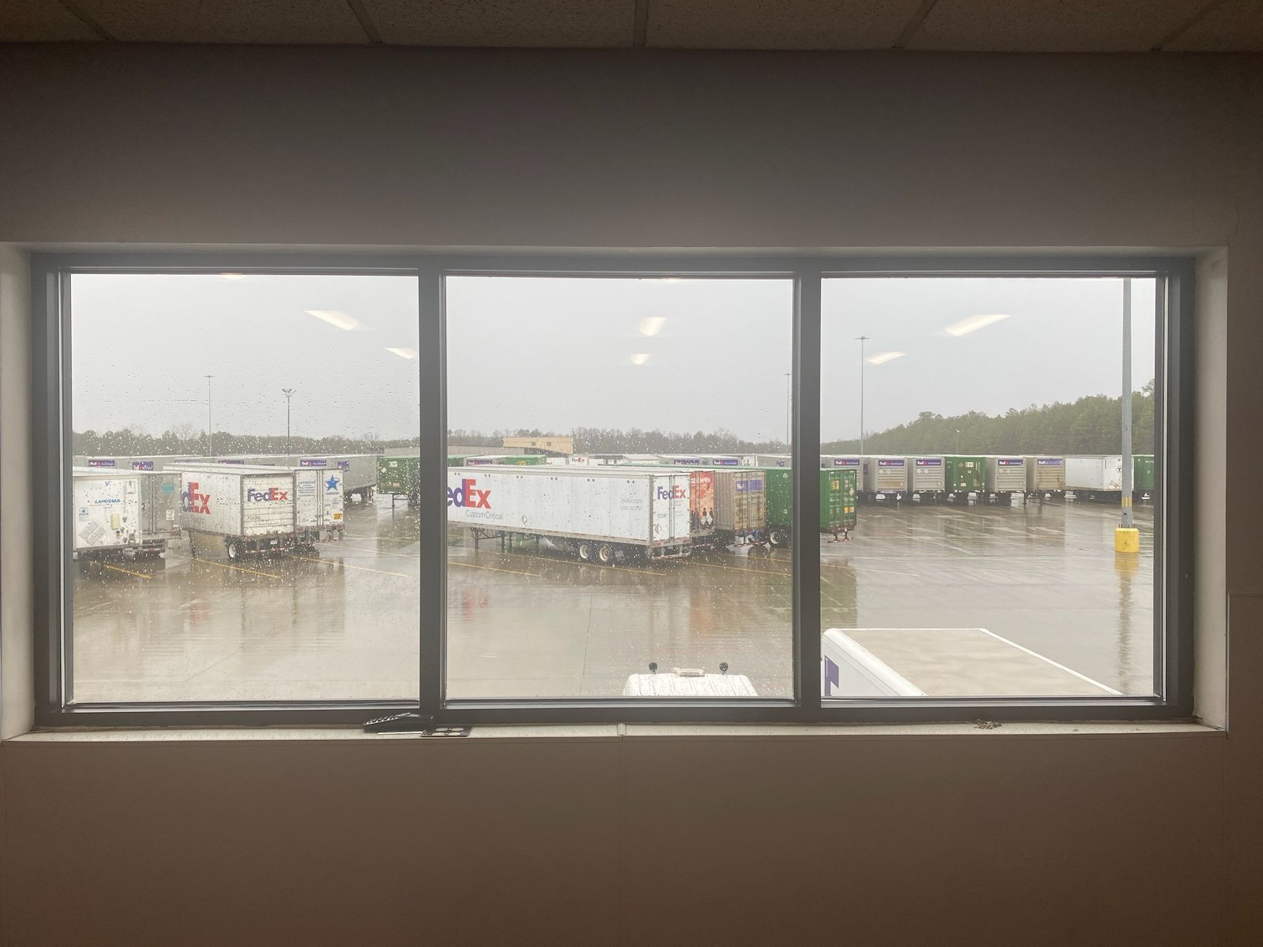 A window with a view of a warehouse with trucks parked in the rain.