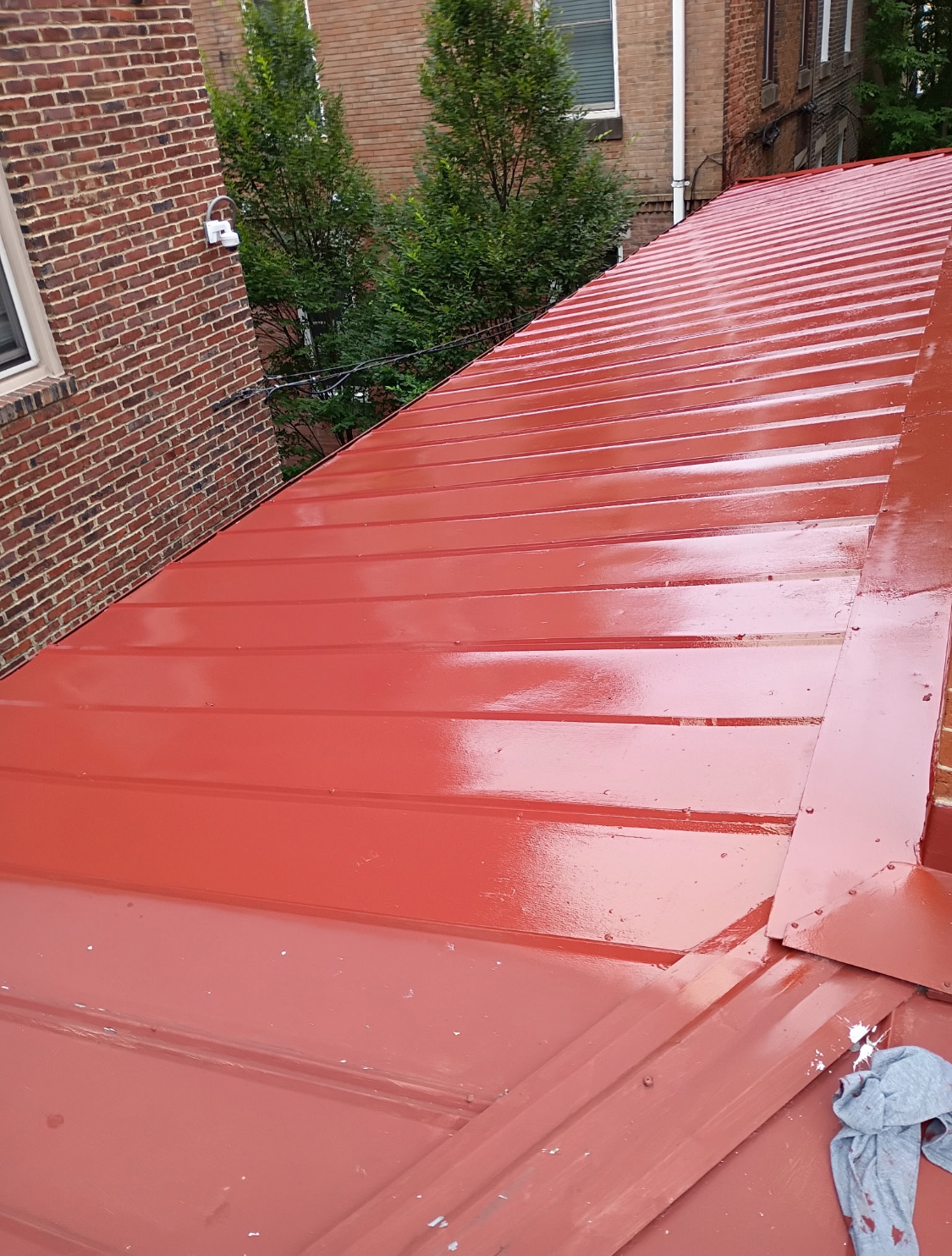 Red metal roof on a building, with trees and a brick wall in the background.