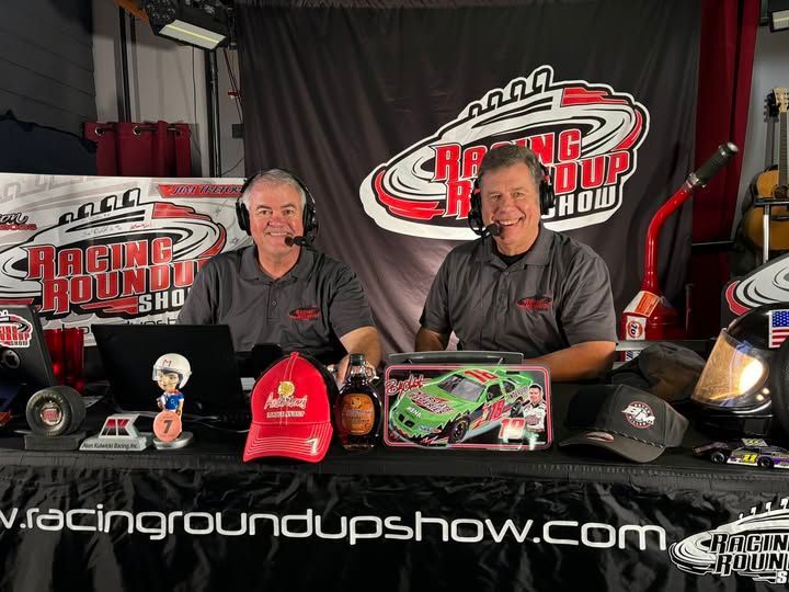 Two hosts behind a Racing Roundup Show table with hats, trophies, and a racing car model on display