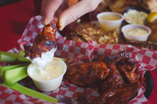 Person dipping a chicken wing in a small cup of white sauce, other wings, celery, and dips in the background.