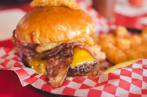 Cheeseburger with bacon, onion ring, and tater tots in red and white checkered basket.