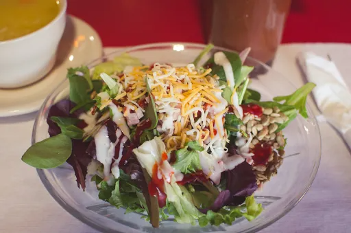 Salad in a glass bowl, topped with shredded cheese and dressing, next to a cup of soup and a drink.