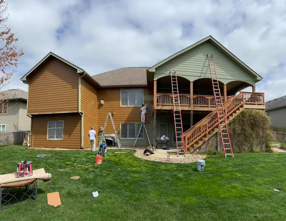 House exterior being painted by multiple workers on ladders; green and brown siding, blue sky.
