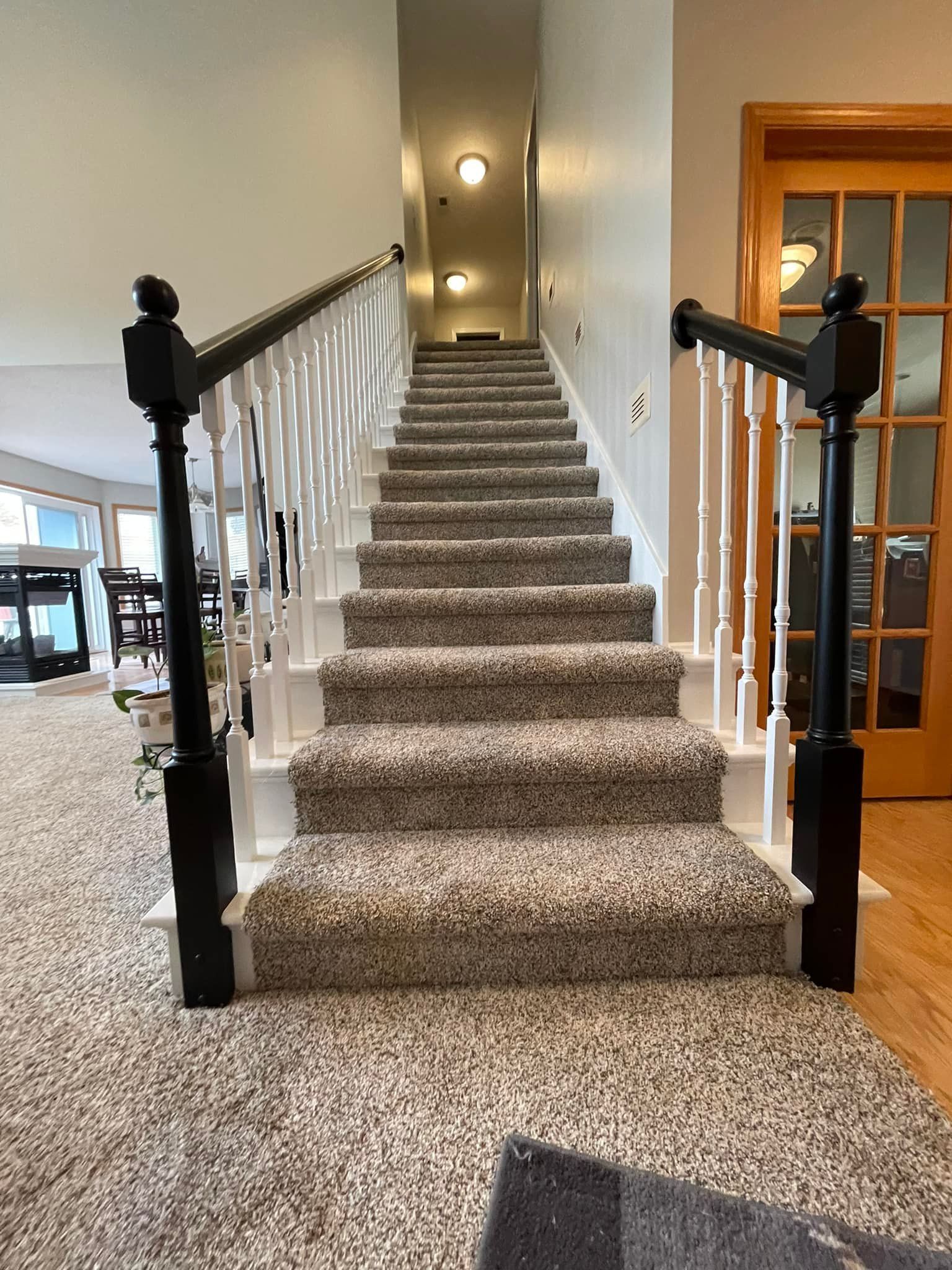 Staircase with carpeted steps, white balusters, black posts, leading upwards. Room with neutral walls and wood door.