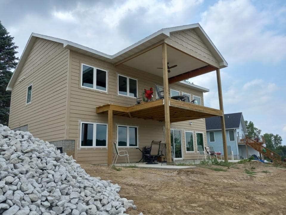 Two-story house with tan siding and a wooden deck, next to a pile of rocks.