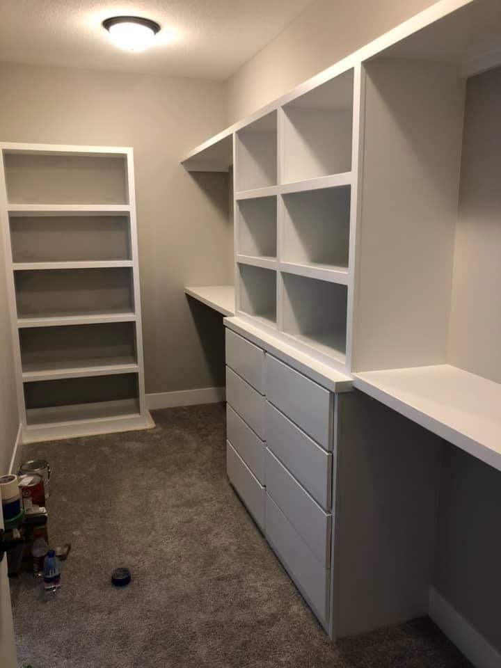 Empty walk-in closet with built-in white shelves, drawers, and countertops. Gray walls, carpet, and a ceiling light.