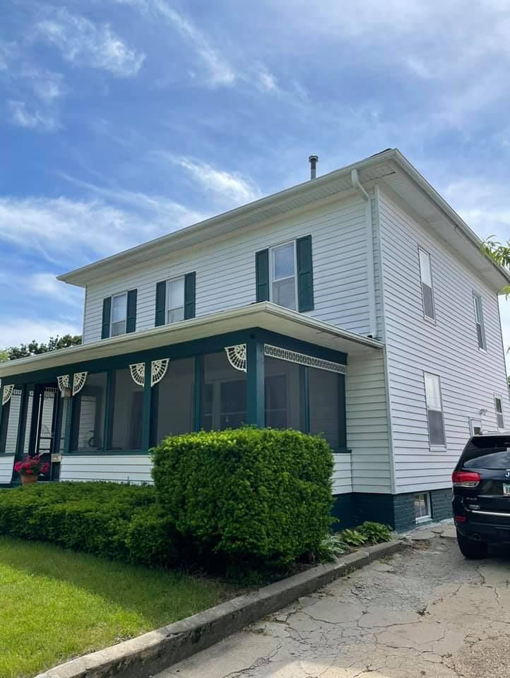Two-story white house with green shutters and screened porch, parked car on right, blue sky.