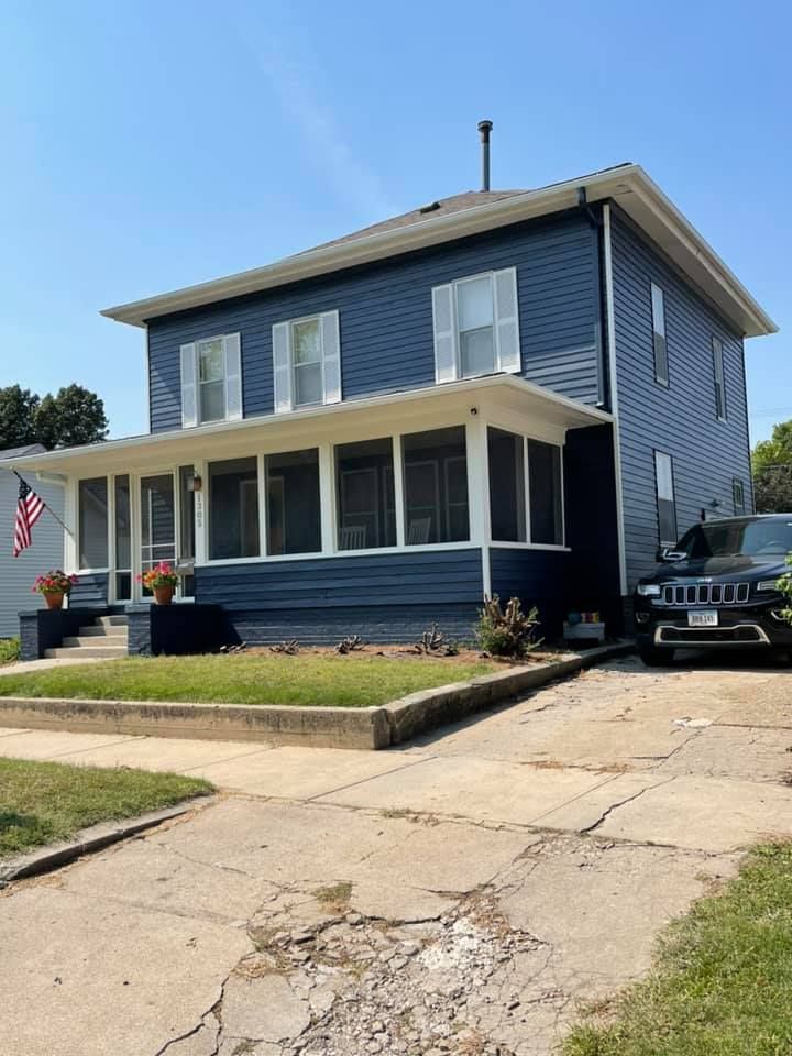 Two-story blue house with porch, white shutters, and driveway. An American flag is visible.