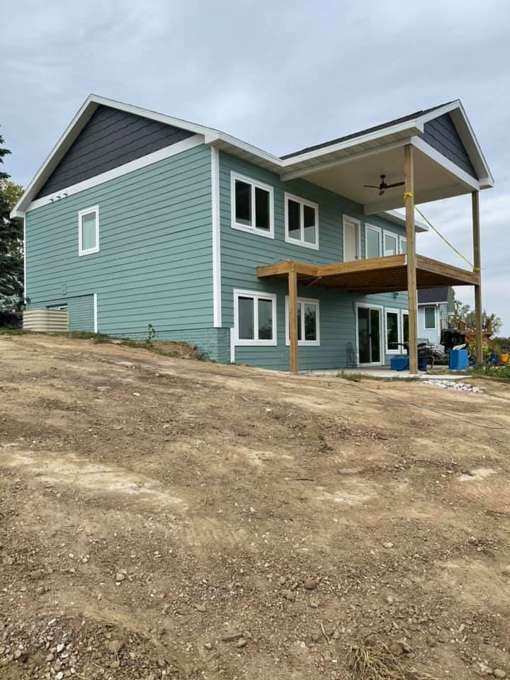 Two-story house with teal siding and a wooden deck, situated on a dirt lot under a cloudy sky.