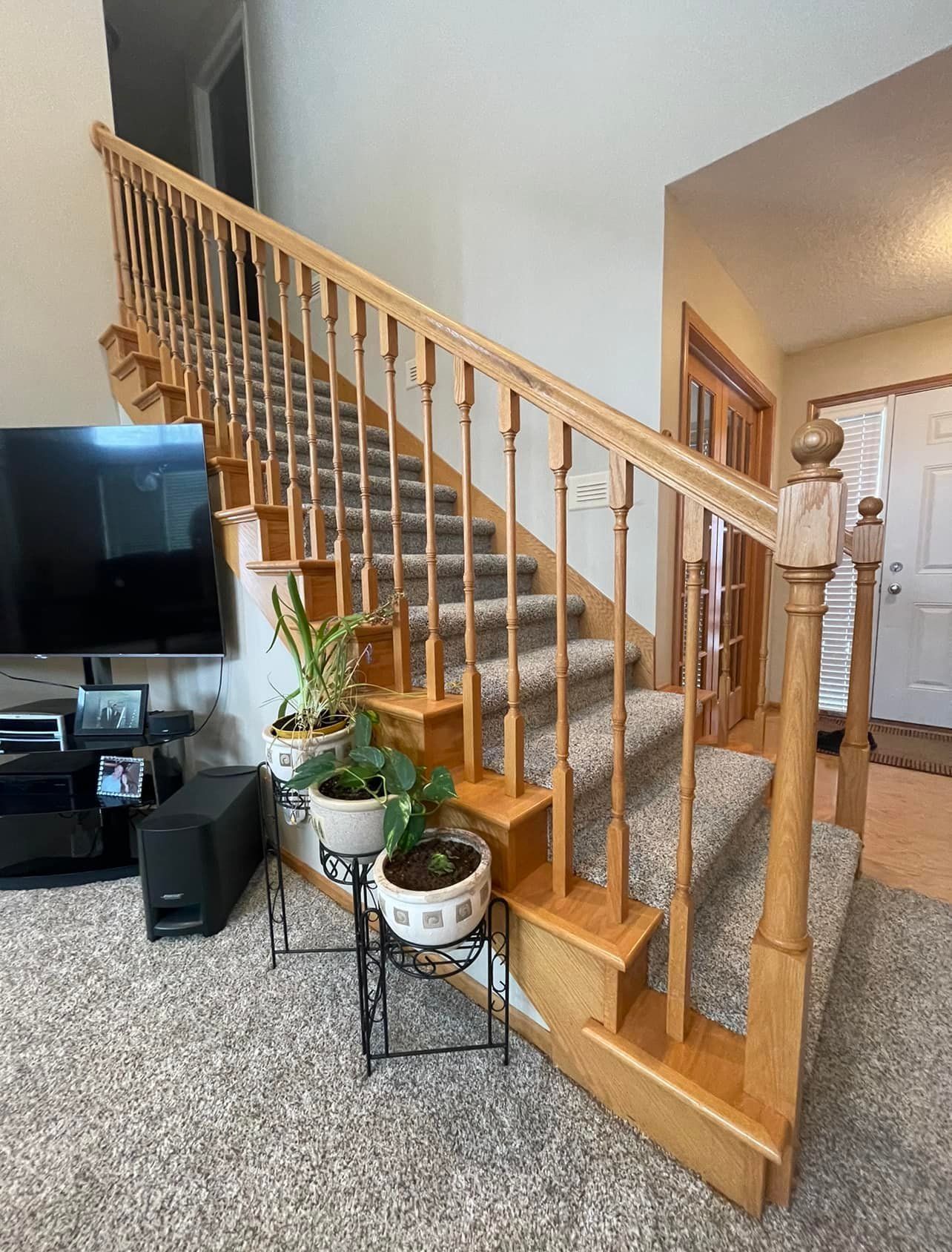Wooden staircase with carpeted steps and a light-colored banister, plants, and a television in a home.
