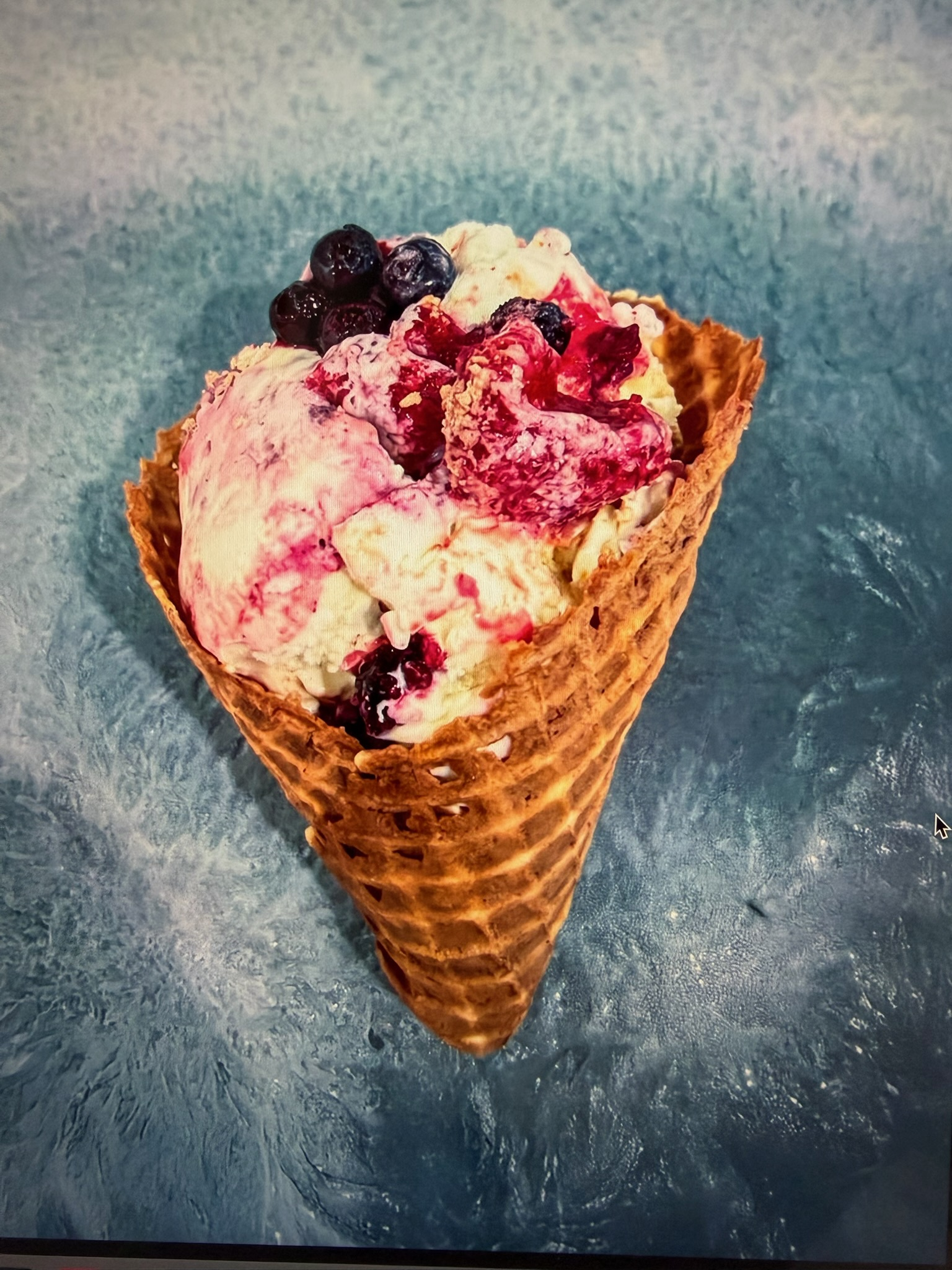 Ice cream cone with pink and white scoops topped with dark berries on a blue surface