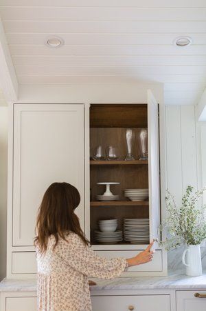 A woman is opening a cabinet in a kitchen filled with plates and glasses.
