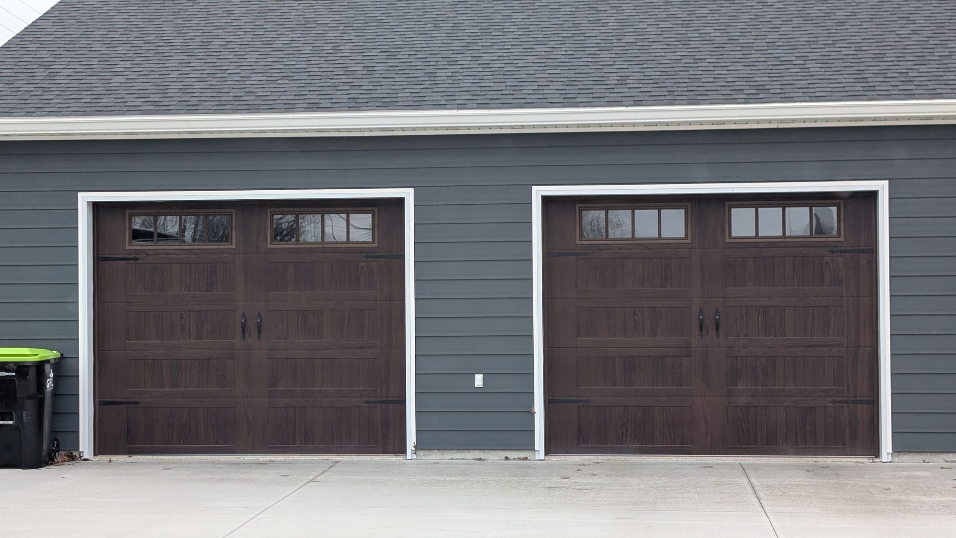 Two dark brown garage doors on a gray building with a concrete driveway in front.