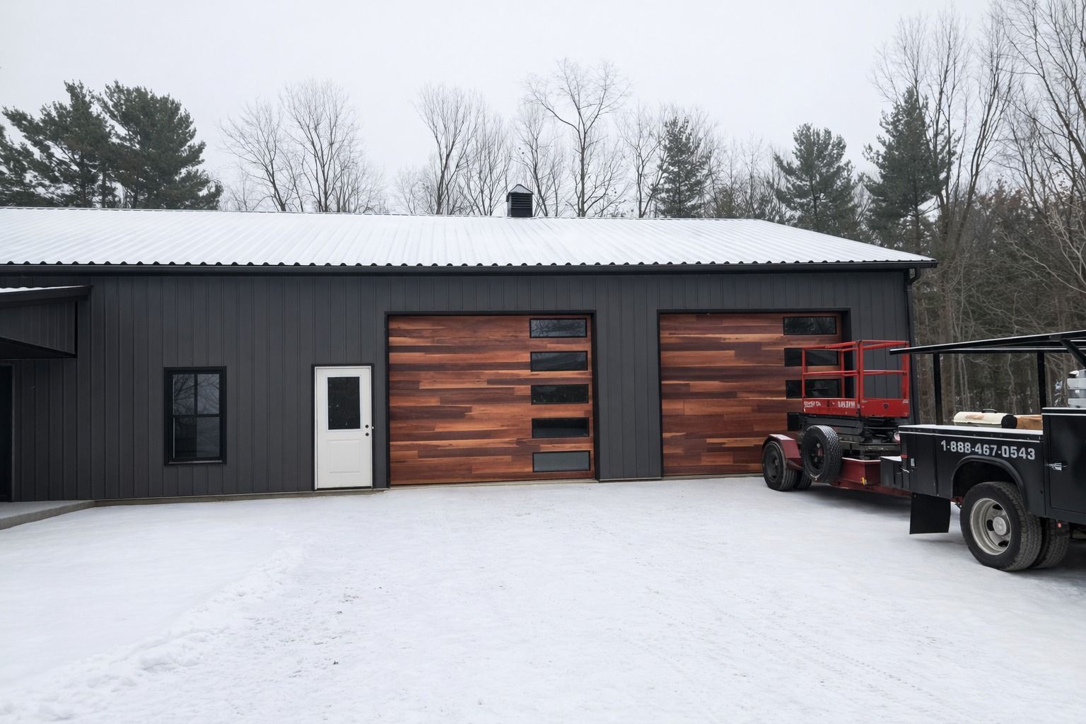 Snow-covered garage with dark siding, wood doors, and a parked utility trailer on the right