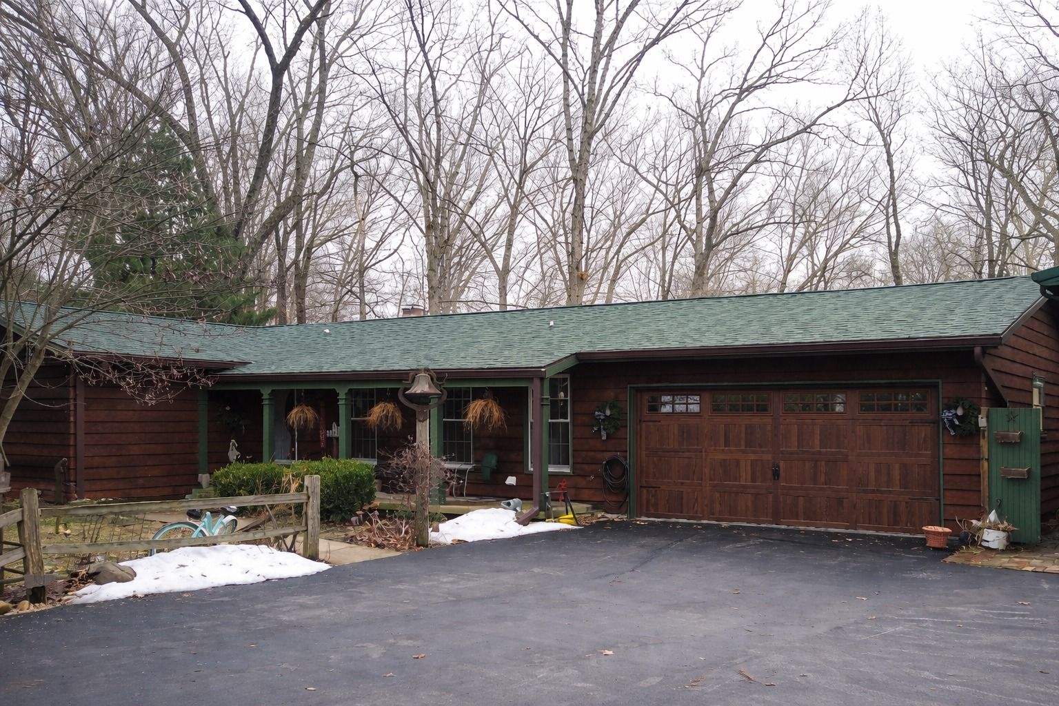 Rustic house with green roof, brown garage, and leafless trees in winter driveway