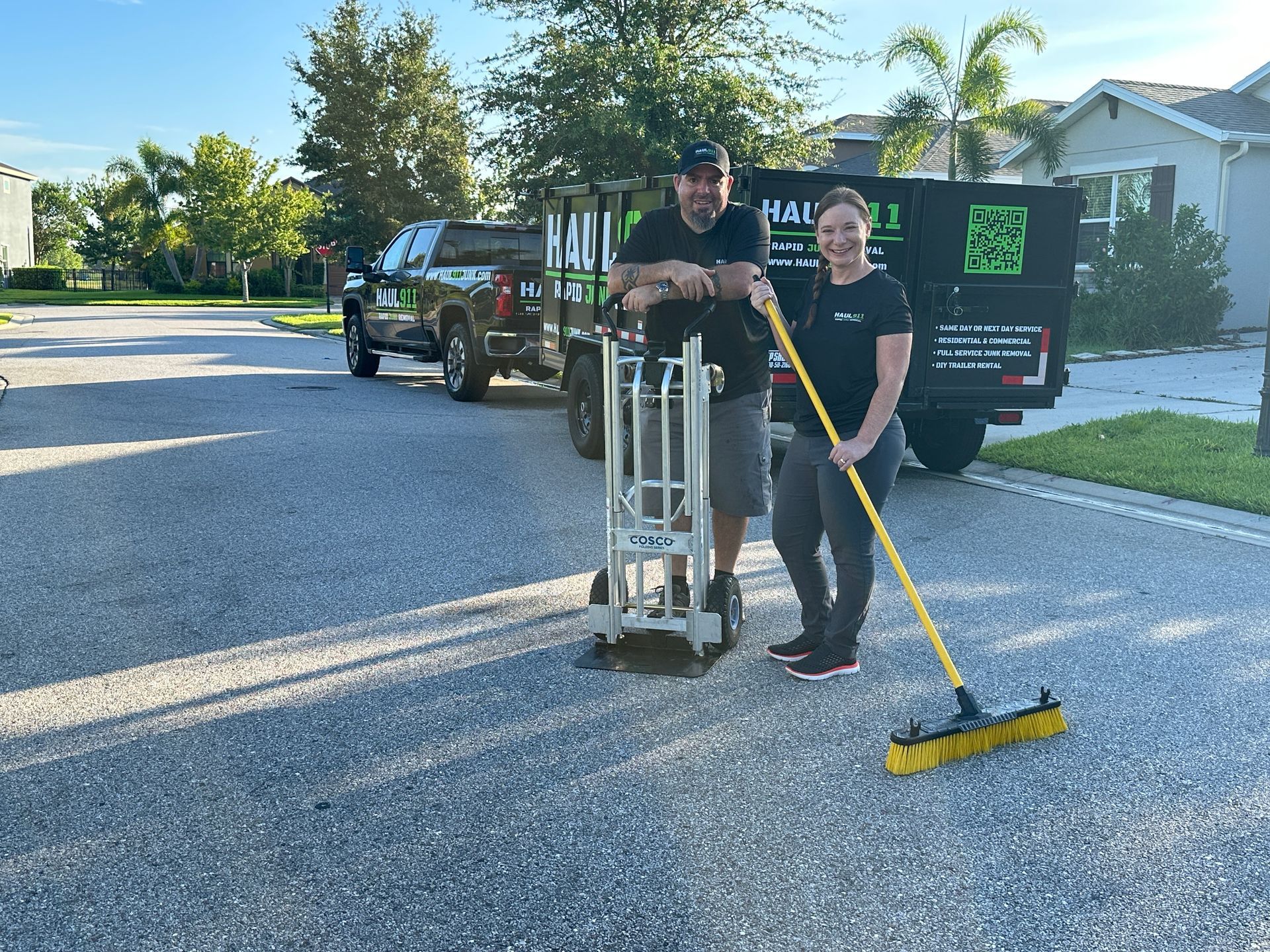 Two people with a truck, hand truck, and broom on a street. Man and woman, both smiling. Truck has
