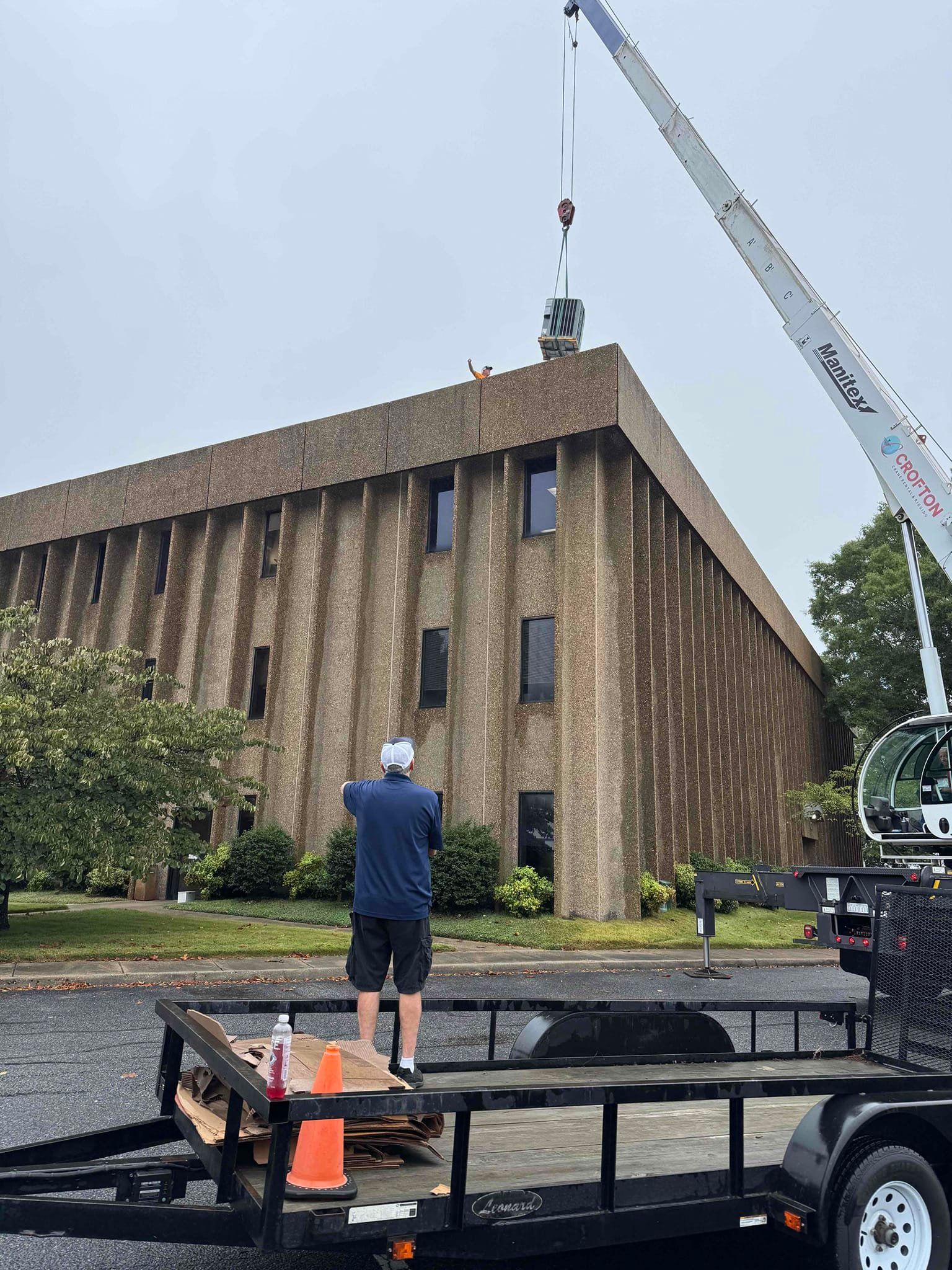 Worker on flatbed trailer near a brick building as a crane lifts equipment overhead.
