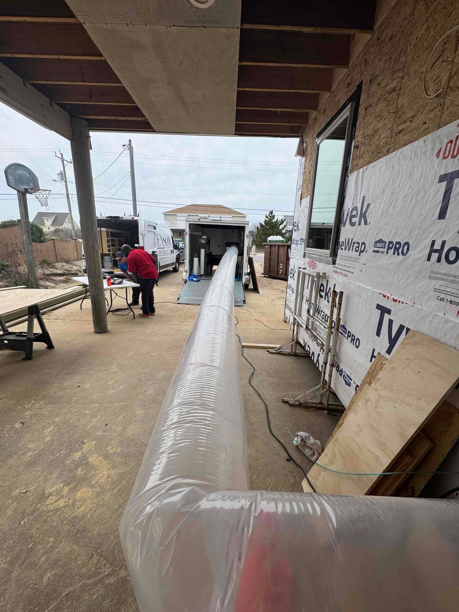 Covered walkway with a long wrapped pipe or duct leading toward a building entrance, with construction materials nearby.