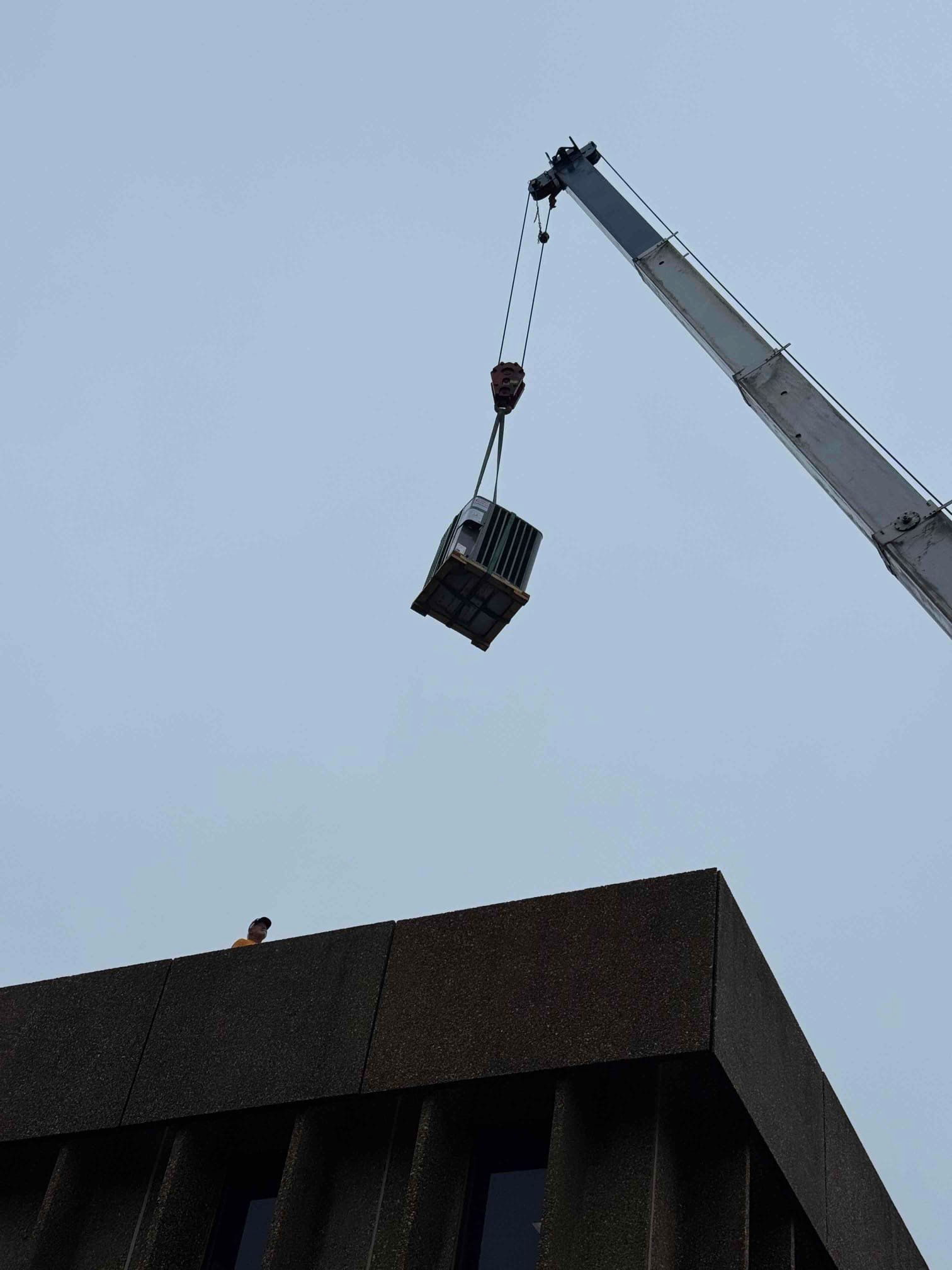 Crane lowering a suspended basket toward a rooftop against a cloudy sky