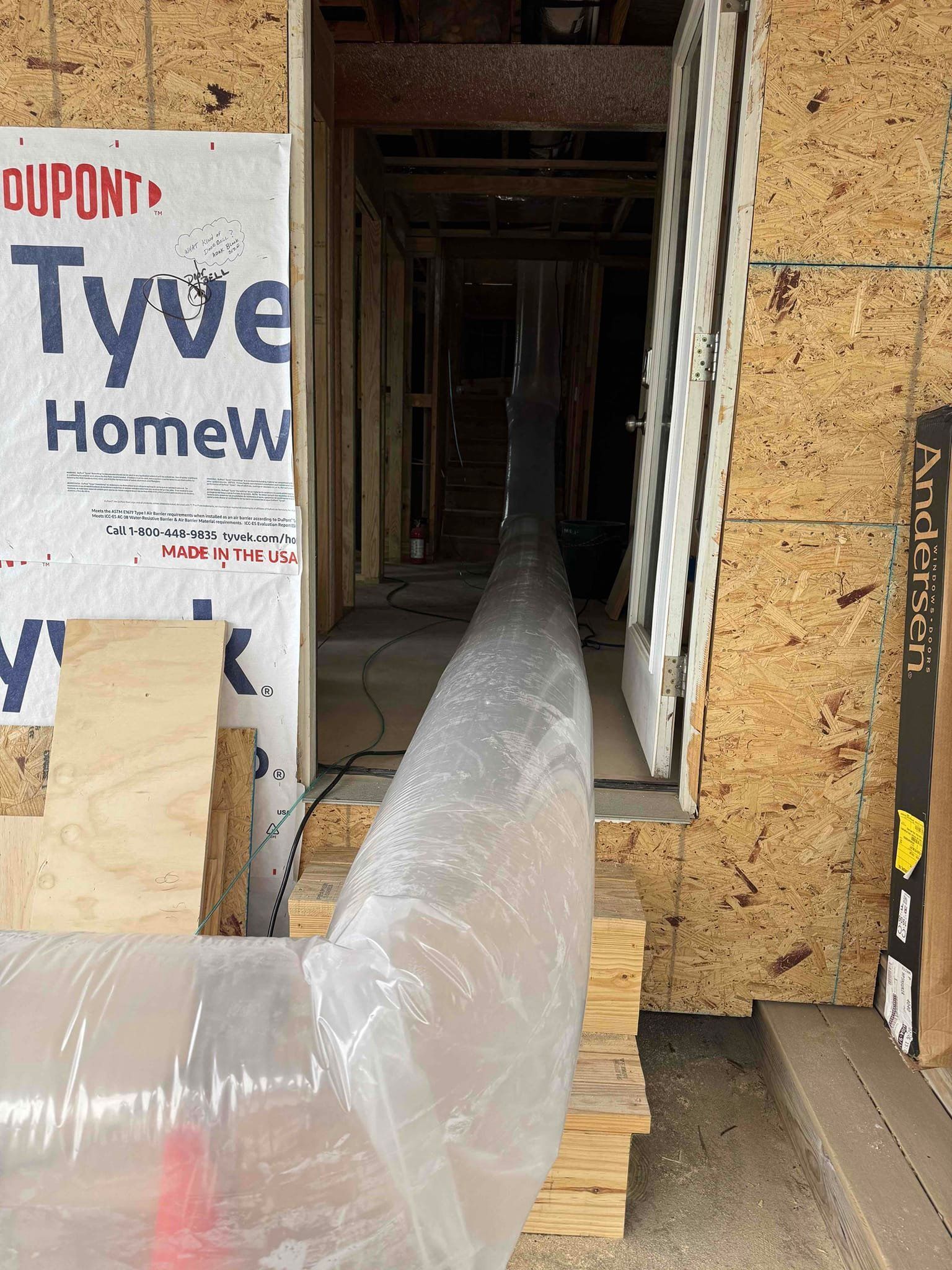 Flexible duct runs through a framed house wall under construction, with insulation and Tyvek wrap visible.