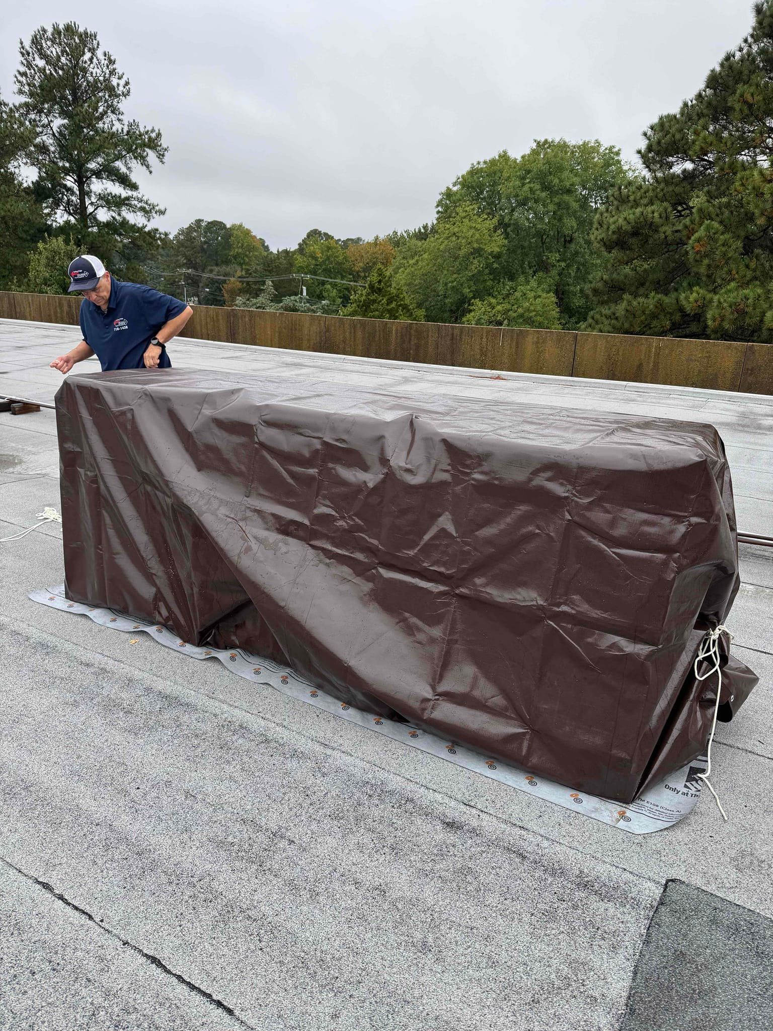 Person crouching beside a large brown tarp-covered object on a gravel rooftop under cloudy skies