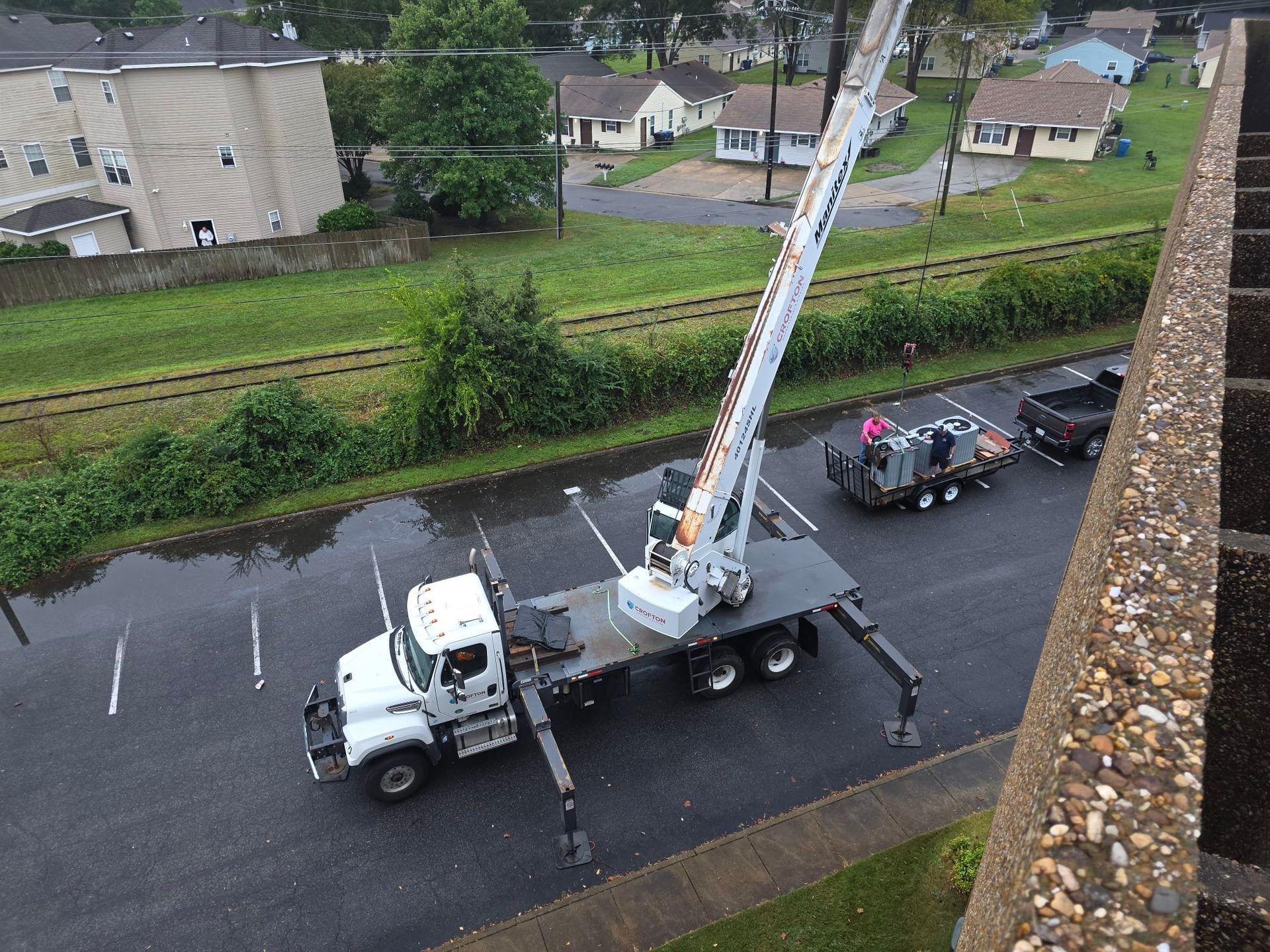White crane truck parked in a lot, lifting gear extended over a dark trailer near railroad tracks and houses