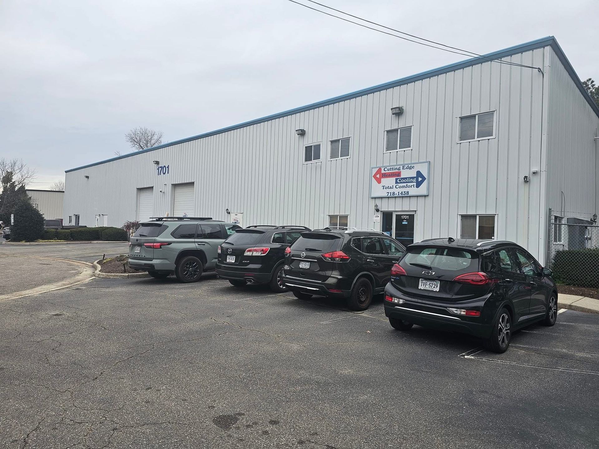 Gray industrial building with a sign, fronted by parked cars on a gravel lot.