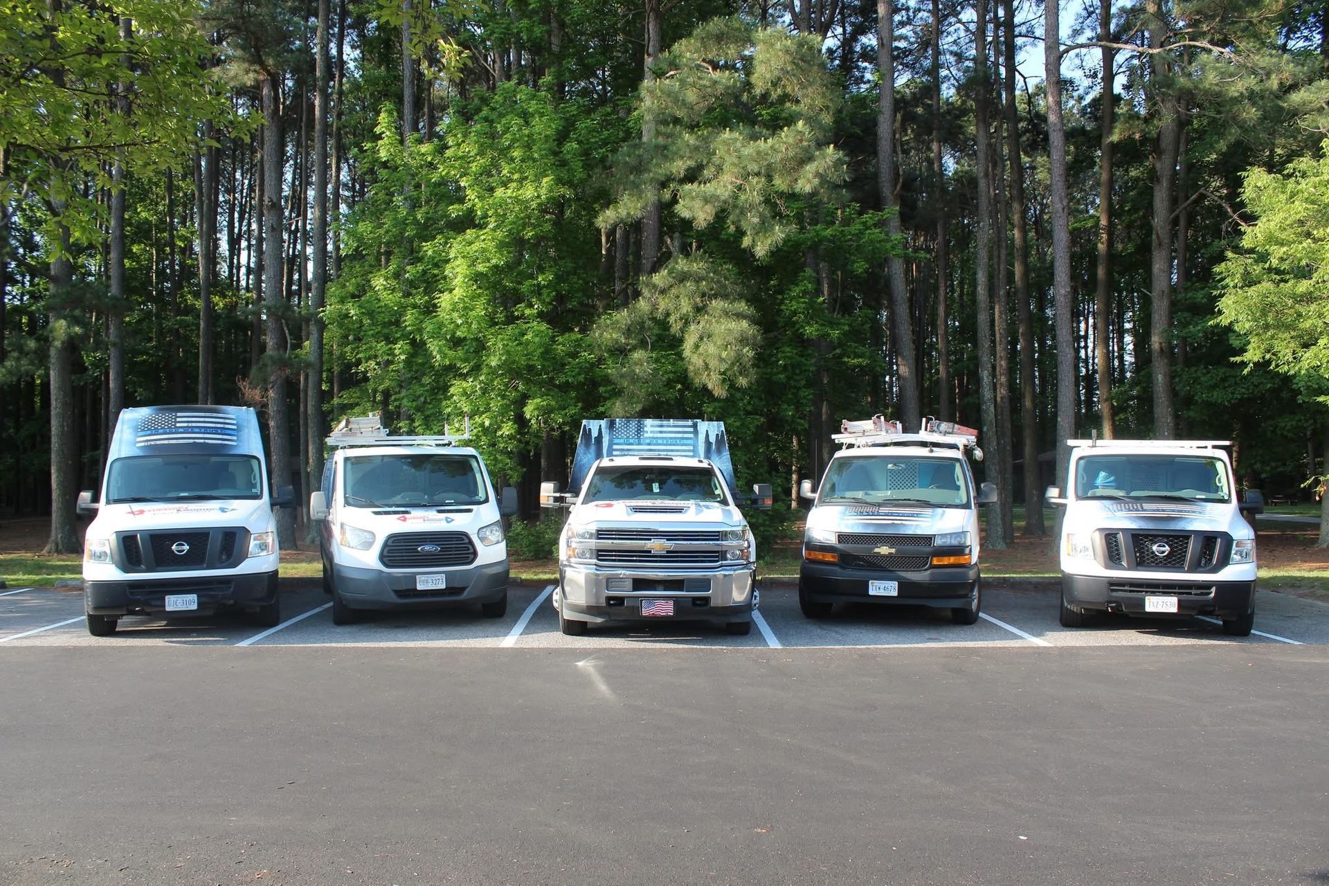 Five white service vans parked side by side in a lot with trees in the background