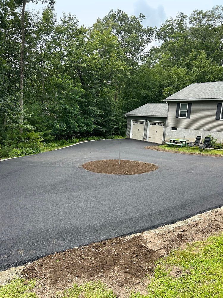 Newly paved asphalt driveway with a circular planting bed in front of a house and garage.
