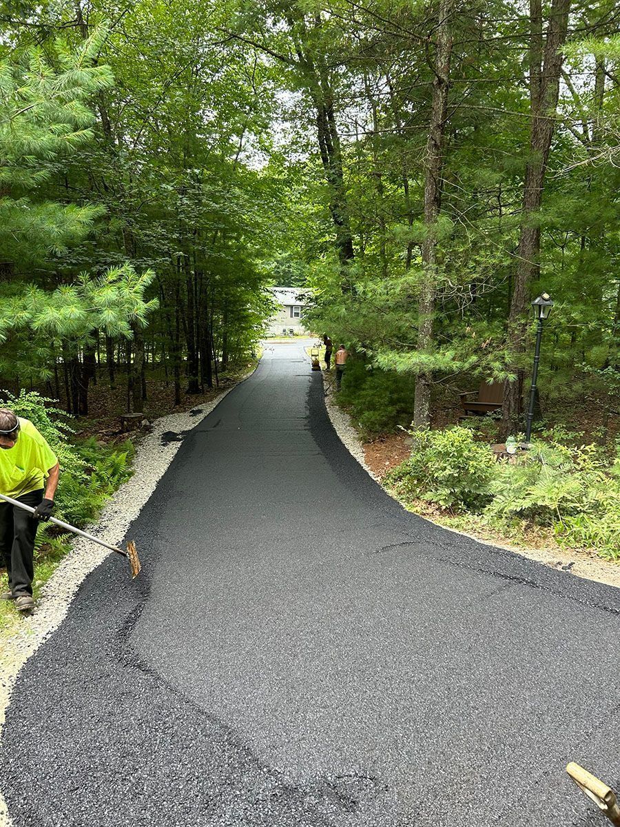 Newly paved asphalt driveway through trees with worker on the left side.