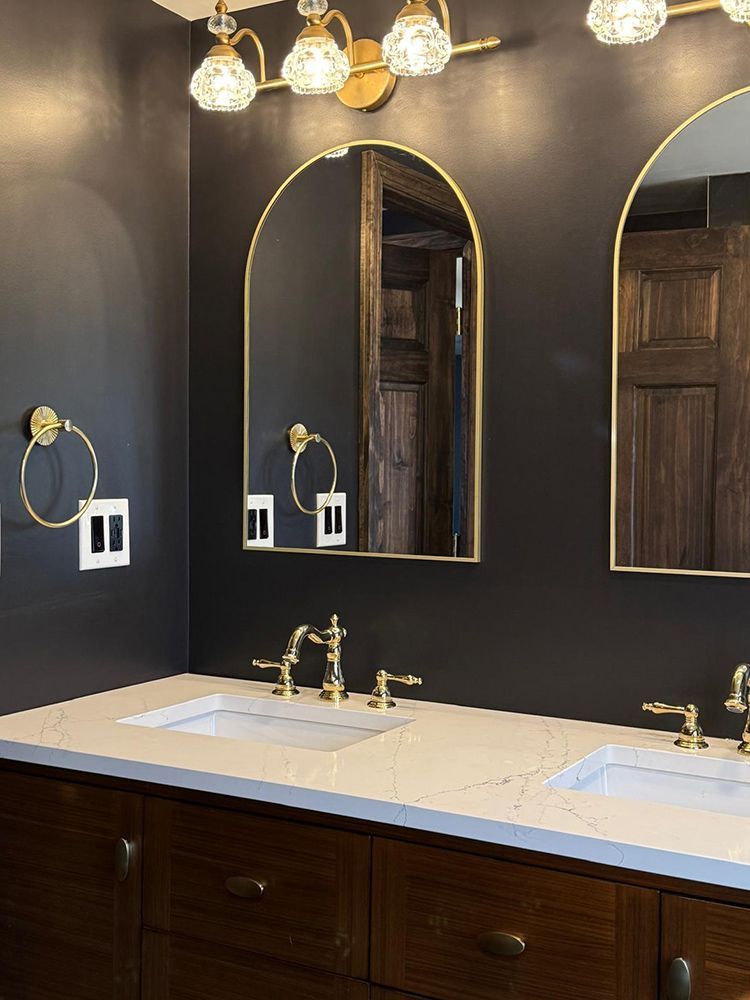 Dark bathroom with gold accents: arched mirrors, light fixture, towel rings, faucets; brown cabinets, white sinks, dark walls.