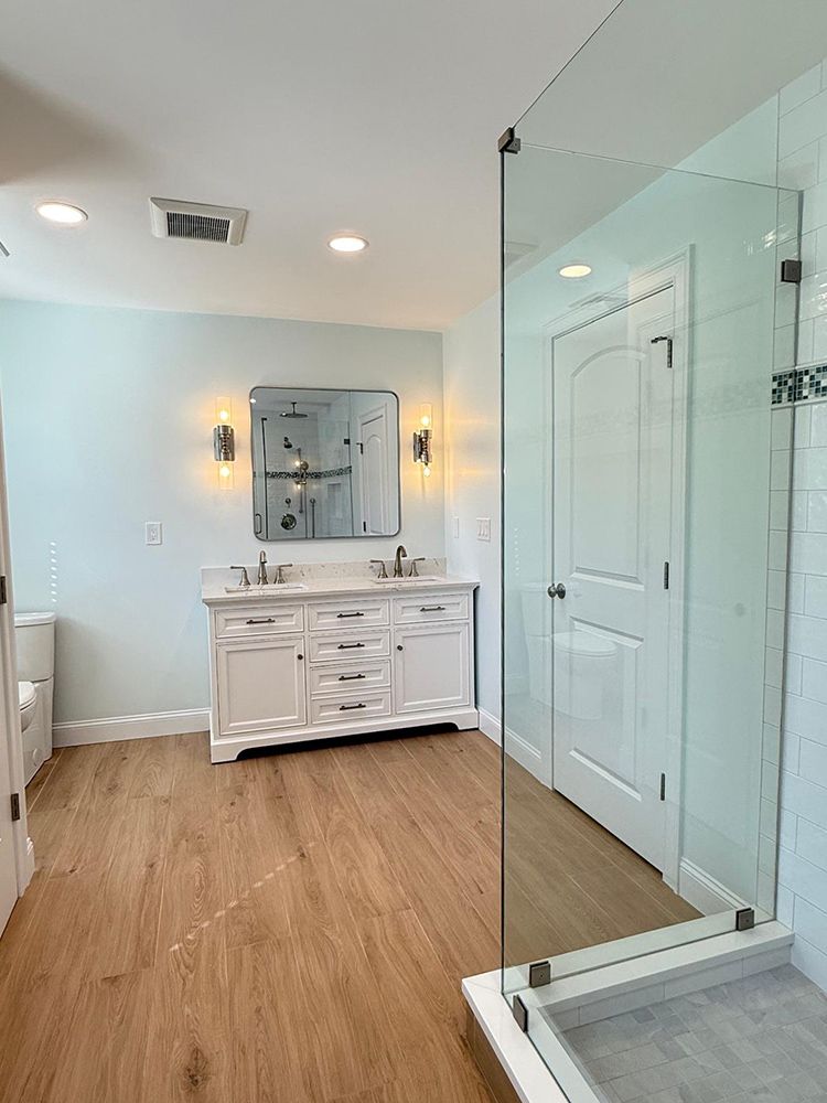 Bathroom with white vanity, light blue walls, glass shower, and wood-look floors.