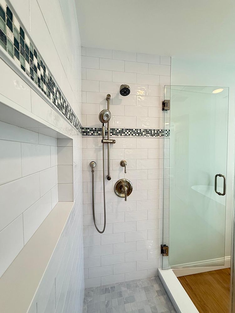 White-tiled shower with glass door, built-in shelf, mosaic tile border, and silver shower fixtures.