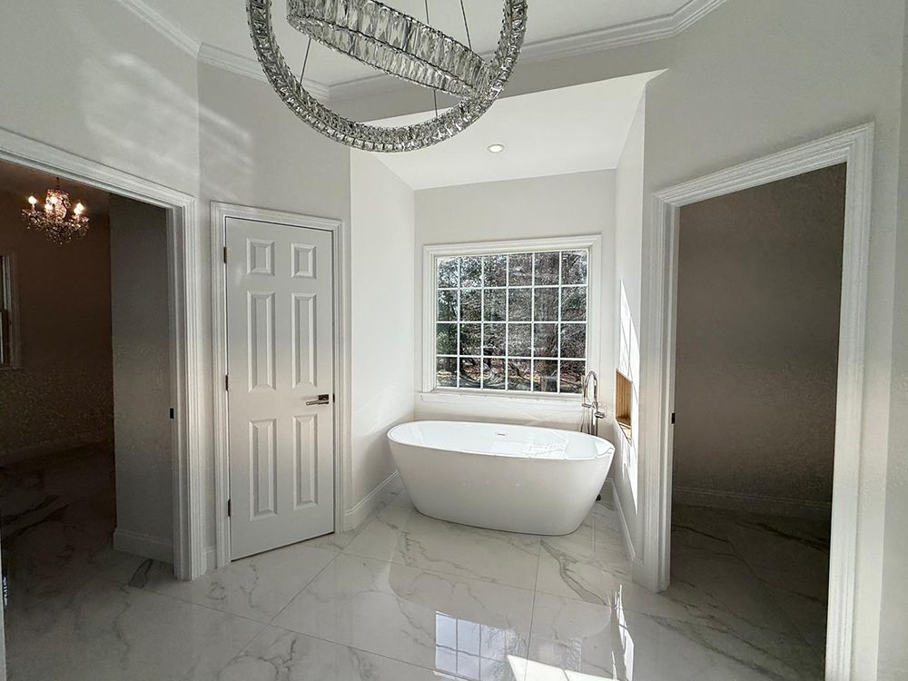 Spacious white bathroom with soaking tub, door, window, and chandelier. Marble floors, light streaming in.