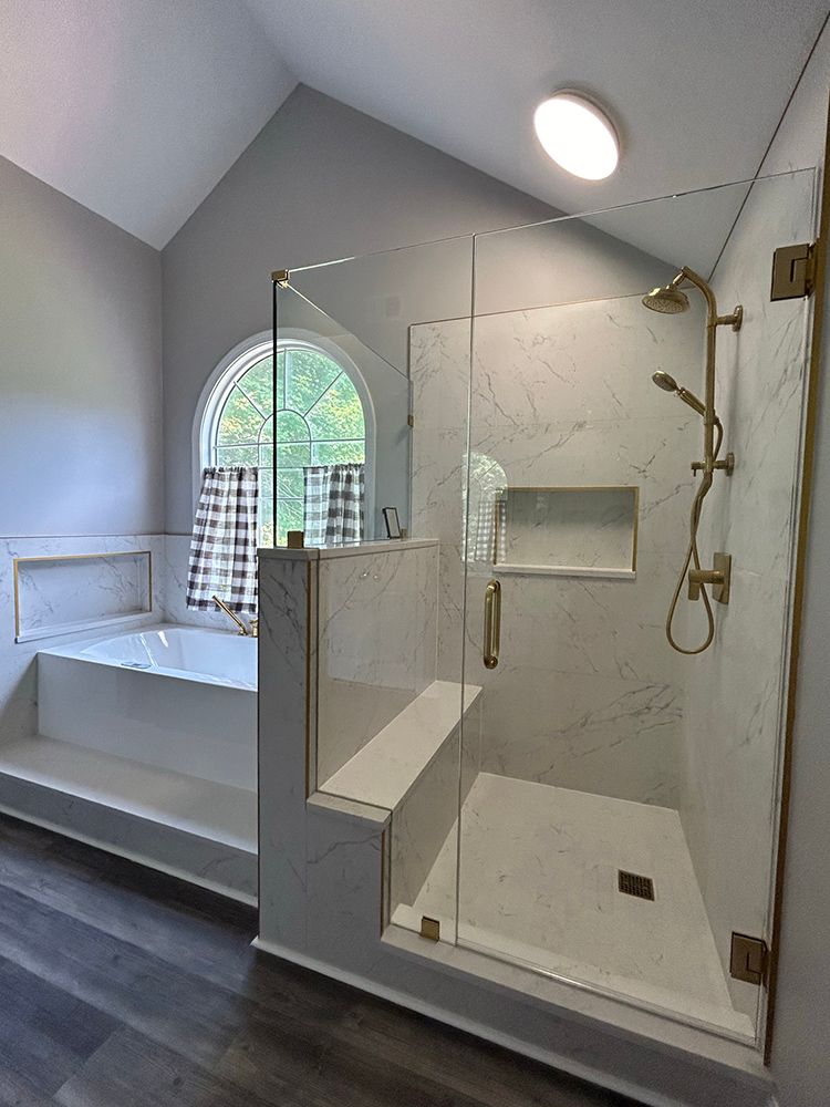 Bathroom with glass shower, gold fixtures, white marble walls, and arched window with plaid curtains.
