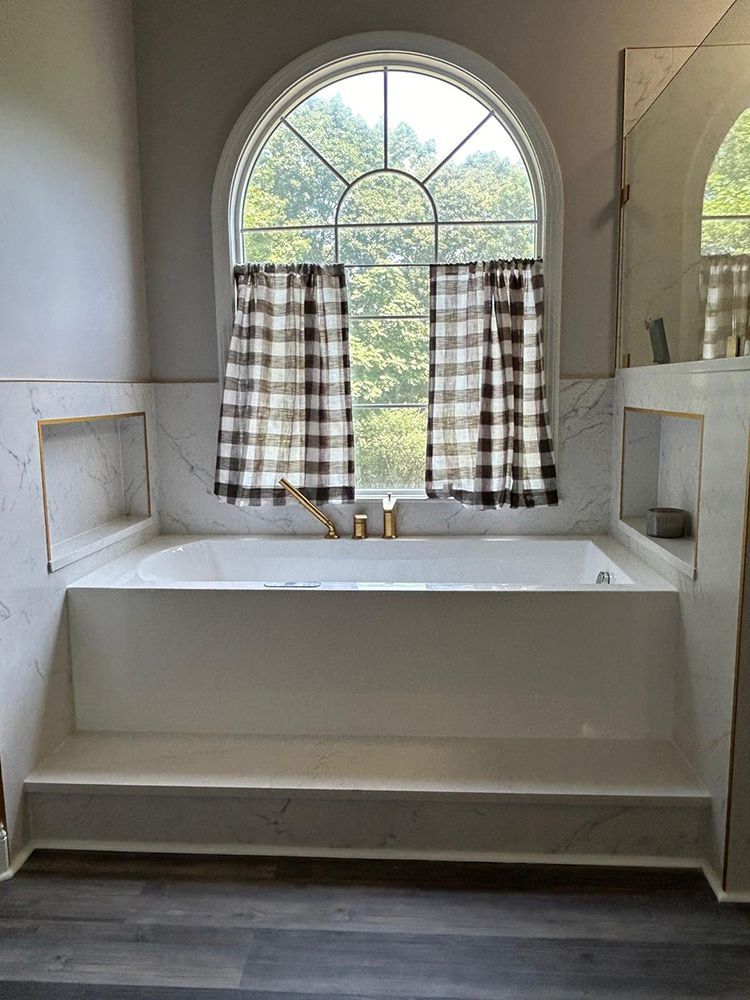 Bathroom with arched window, bathtub, plaid curtains, and gray and white marble accents.