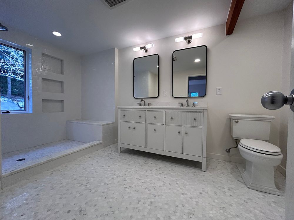 White bathroom with double vanity, mirrors, and toilet. Mosaic tile floor, recessed shelves, and walk-in shower.