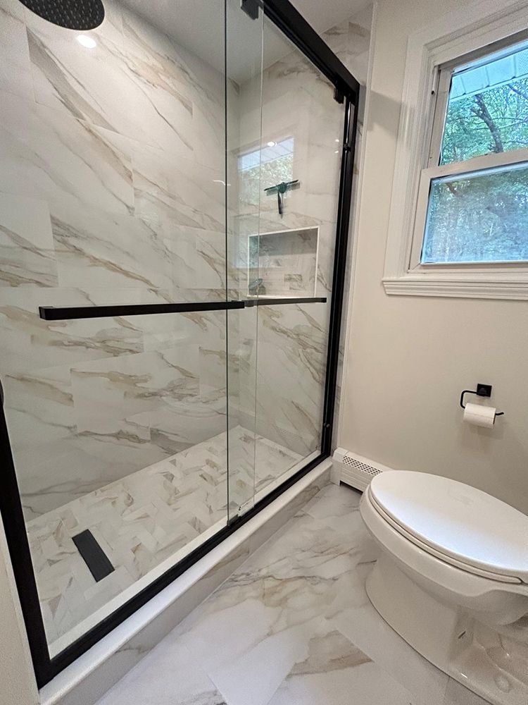 Modern bathroom with black-framed shower, marbled tile, toilet, and window.