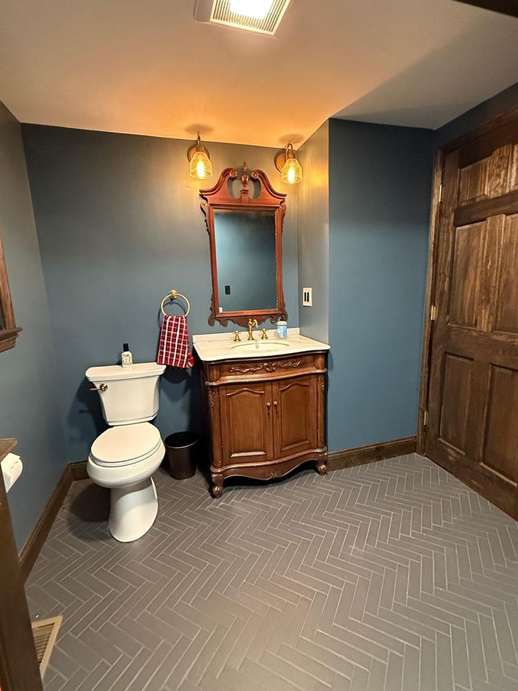 Small bathroom with blue walls, wooden vanity, ornate mirror, and herringbone tile floor.