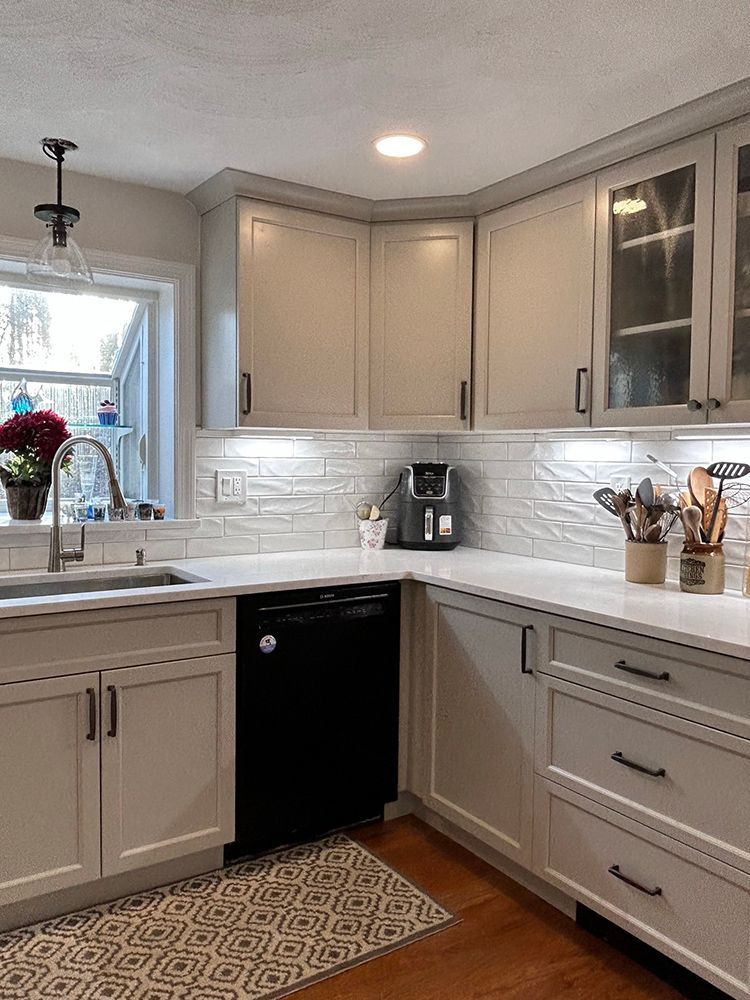 A kitchen with light gray cabinets, white countertops, a black dishwasher, and a window with a sink.