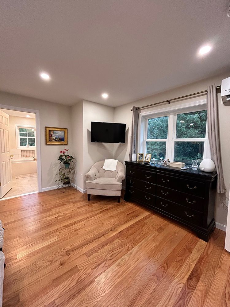 Bedroom with hardwood floor, beige walls, black dresser, armchair, and window with greenery.