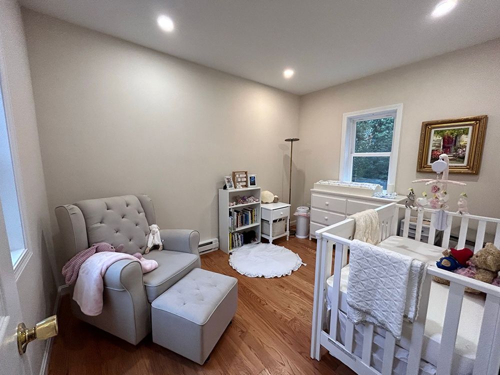 Nursery with a crib, glider, dresser, and rug; tan walls, wood floor, and window with a picture.