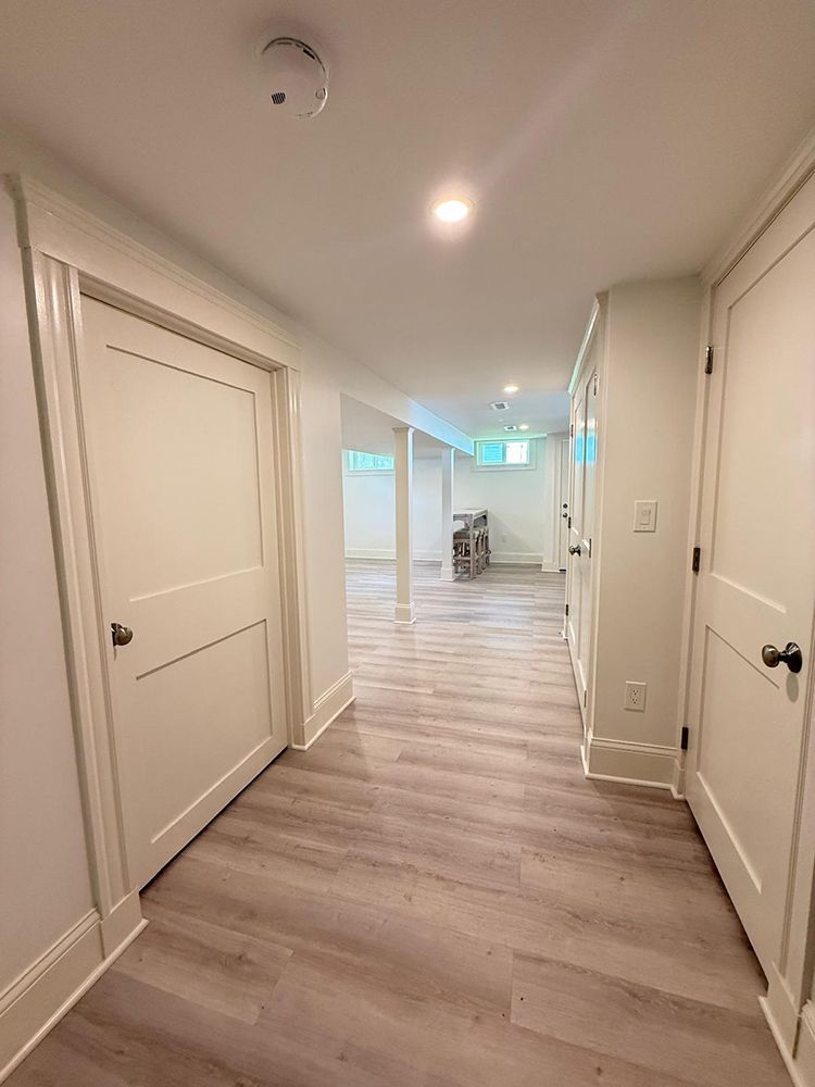 Narrow hallway with white doors, gray wood-look flooring, white walls, and recessed lighting.
