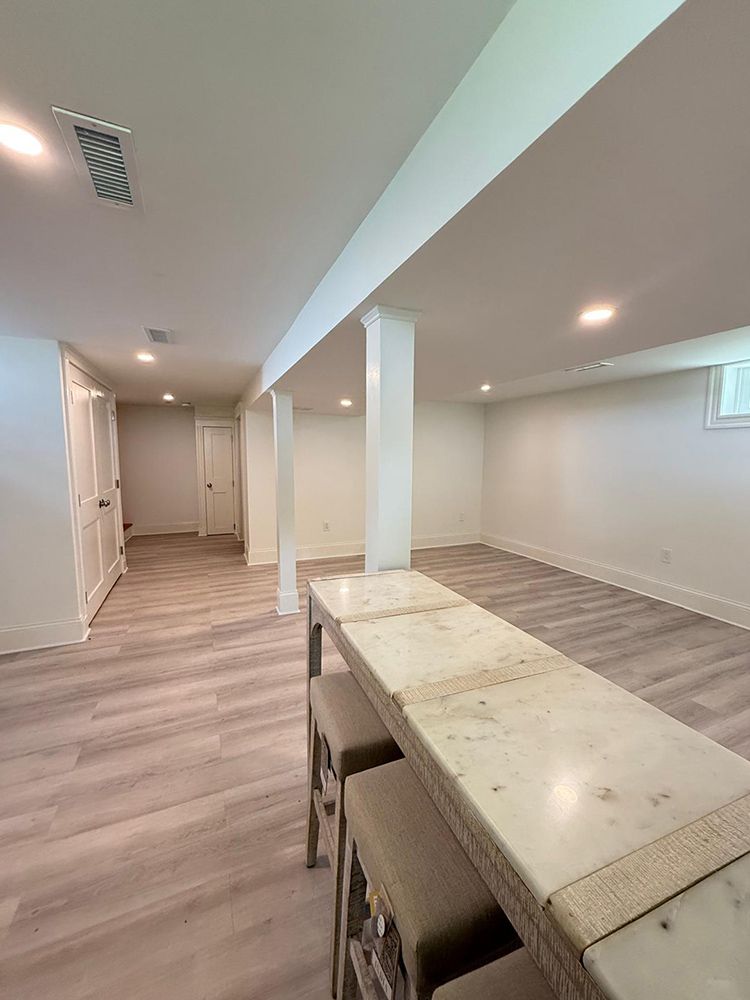Spacious, white-walled basement with a marble-topped bar and stools, light wood-look floors, and recessed lighting.